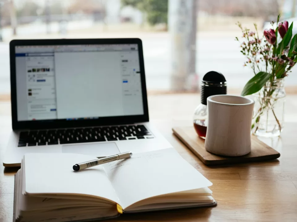 Laptop open on a white desk beside a coffee cup, notebook and pen